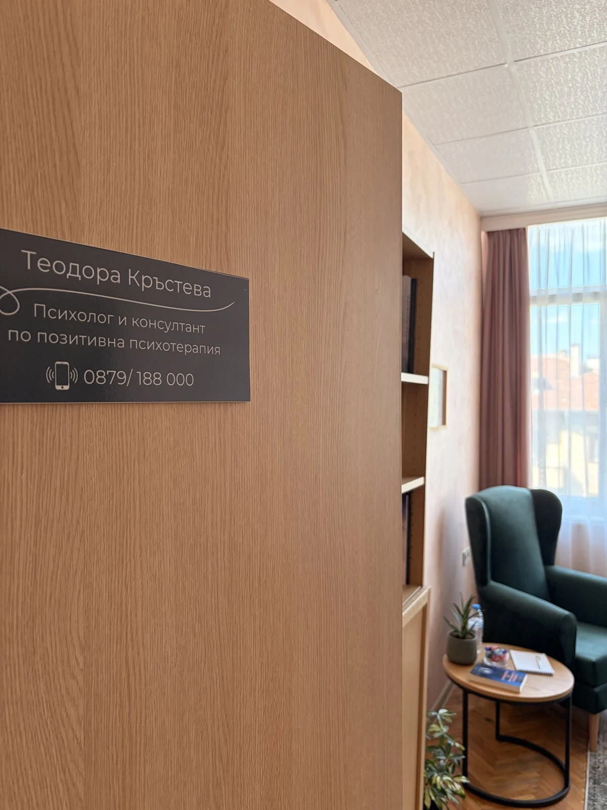 Отворената врата на кабинета с табелата „Теодора Кръстева, психолог и консултант по позитивна психотерапия“ и зеленото кресло до прозореца